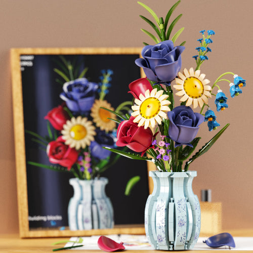 Colorful artificial flowers in a vase on a desk with a framed picture of flowers in the background.
